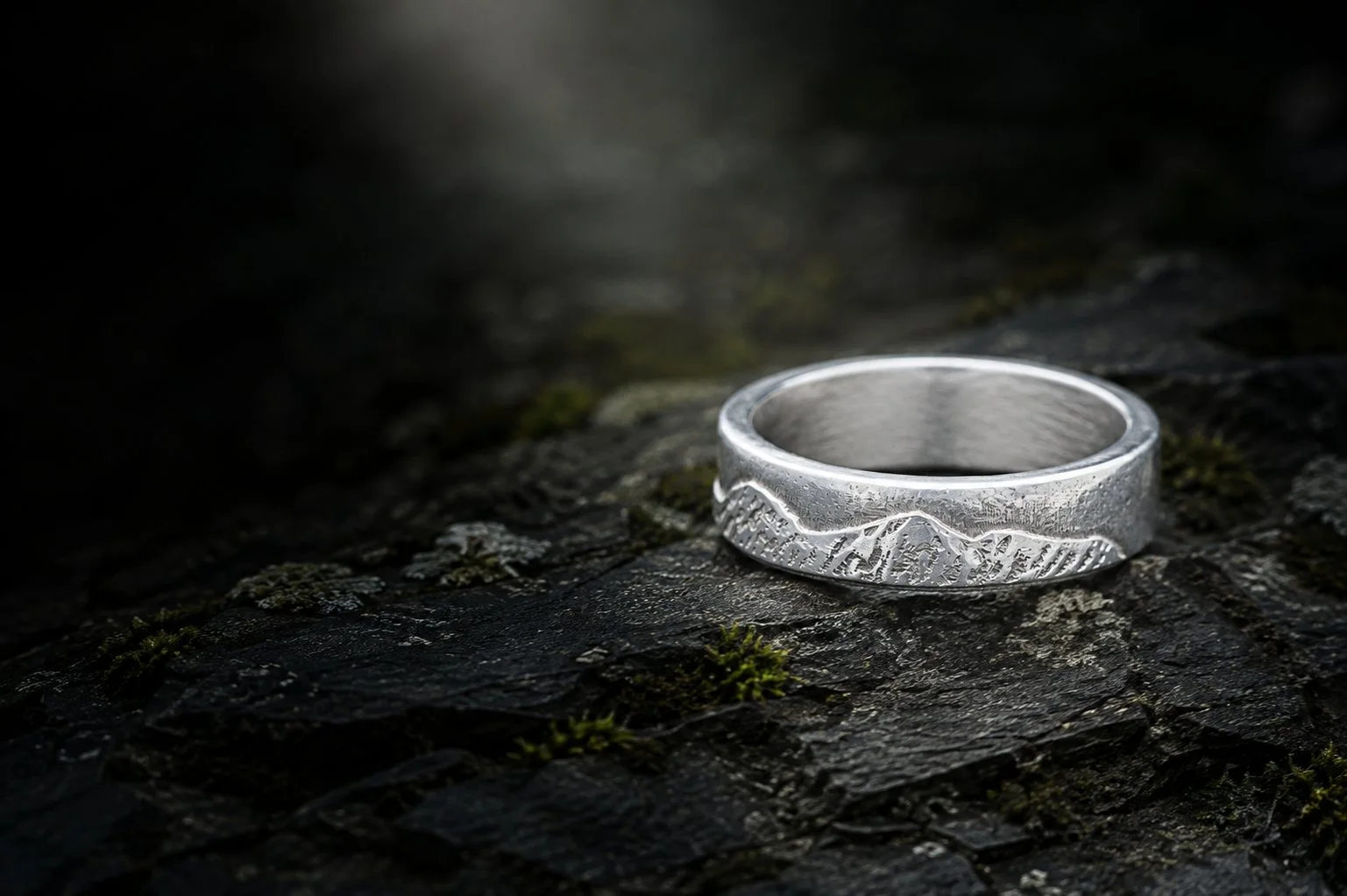 The National Three Peaks silver ring resting on dark weathered slate among moss, in low evening light.